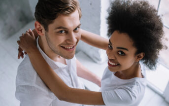 A smiling couple stands close together, looking up at the camera. They have their arms around each other. The man has short, wavy hair and is wearing a white shirt. The woman has curly hair and is also wearing a white shirt. Bright light from a window illuminates them.