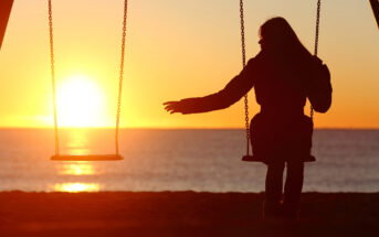 Silhouetted person sitting on a swing at the beach, extending a hand towards another swing next to them, with a vibrant sunset over the ocean in the background. The golden light of the setting sun creates a serene and reflective atmosphere.