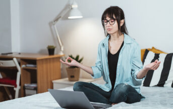 A person with glasses sits cross-legged on a bed, meditating with their eyes closed and hands in a mudra position. A laptop is open in front of them. The background includes a desk, a chair, a lamp, and a potted plant.