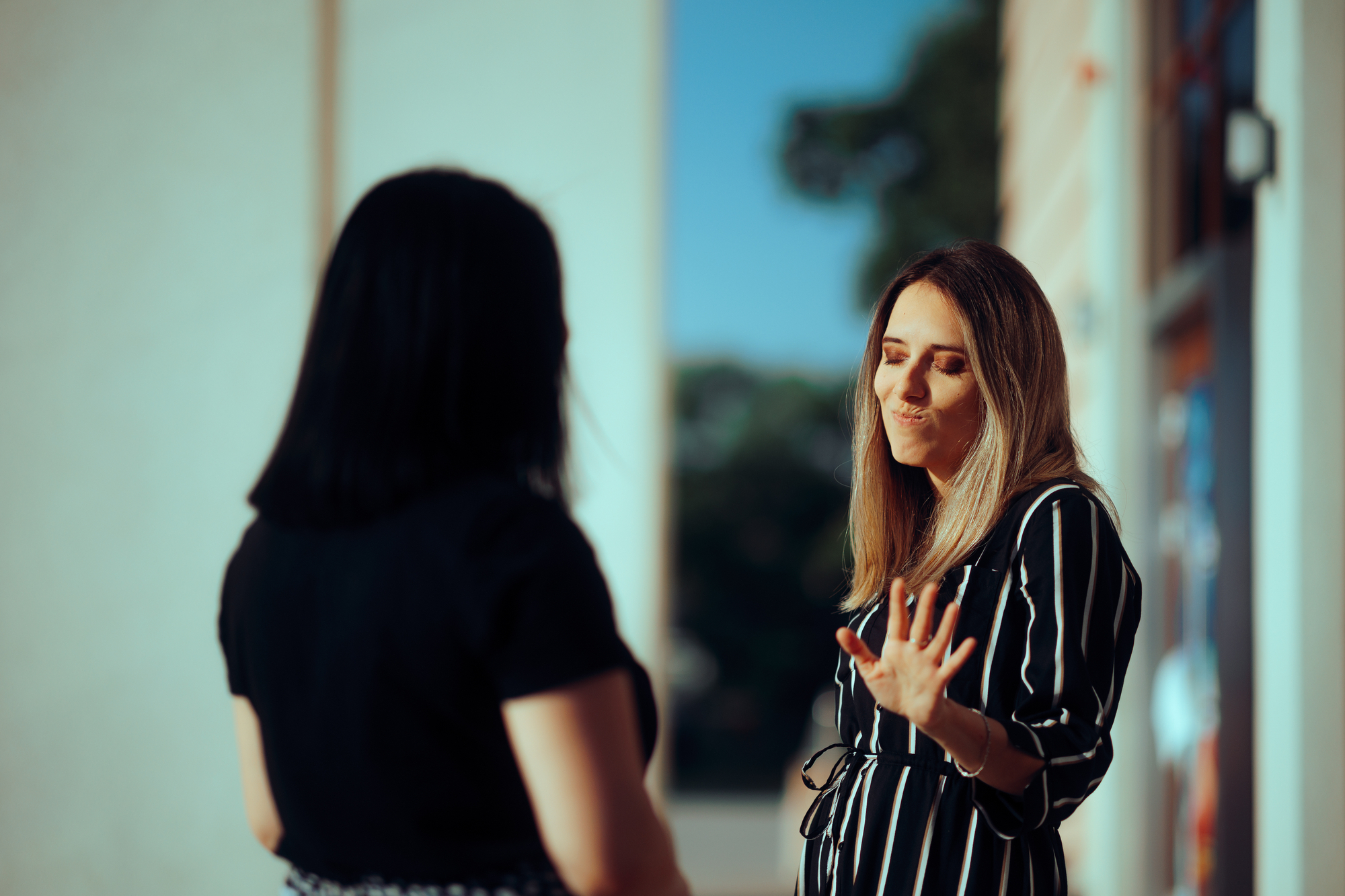 Two women are standing outside in conversation. The woman on the right, wearing a black and white striped top, has her eyes closed and hand raised as if making a point. The woman on the left, with dark hair, is facing away from the camera.