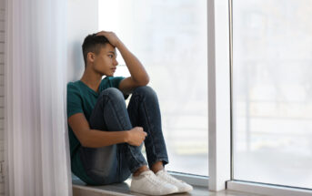 A young person sits on a windowsill, staring thoughtfully outside. They have short hair, wear a green t-shirt, jeans, and white sneakers. One arm rests on their knee, while the other hand touches their head. Light pours in from the large window beside them.