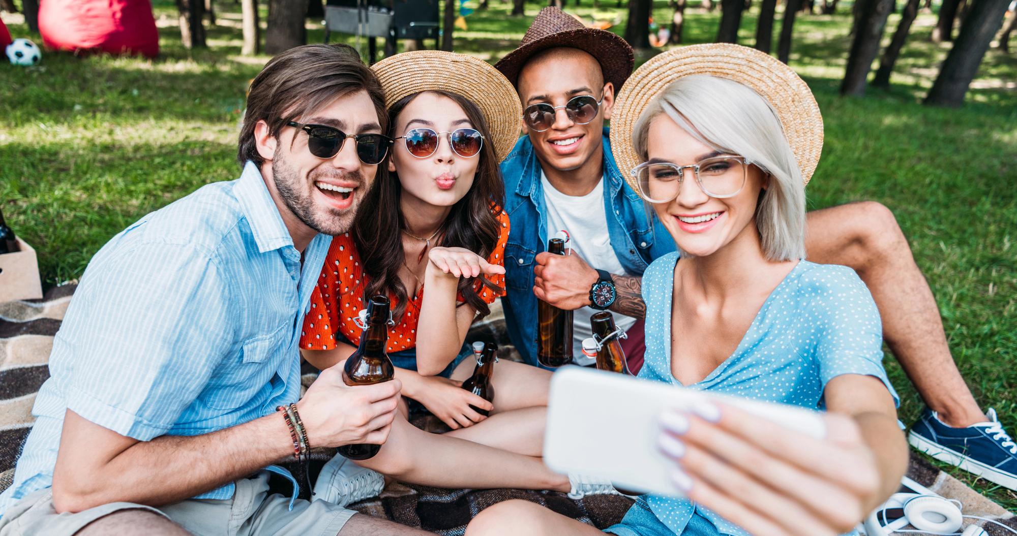 A group of four friends sits on a blanket outdoors, smiling and posing for a selfie. Two wear sunglasses and hats, while one blows a kiss to the camera. They are all dressed casually, enjoying a sunny day with drinks in hand. Trees and grass are visible in the background.