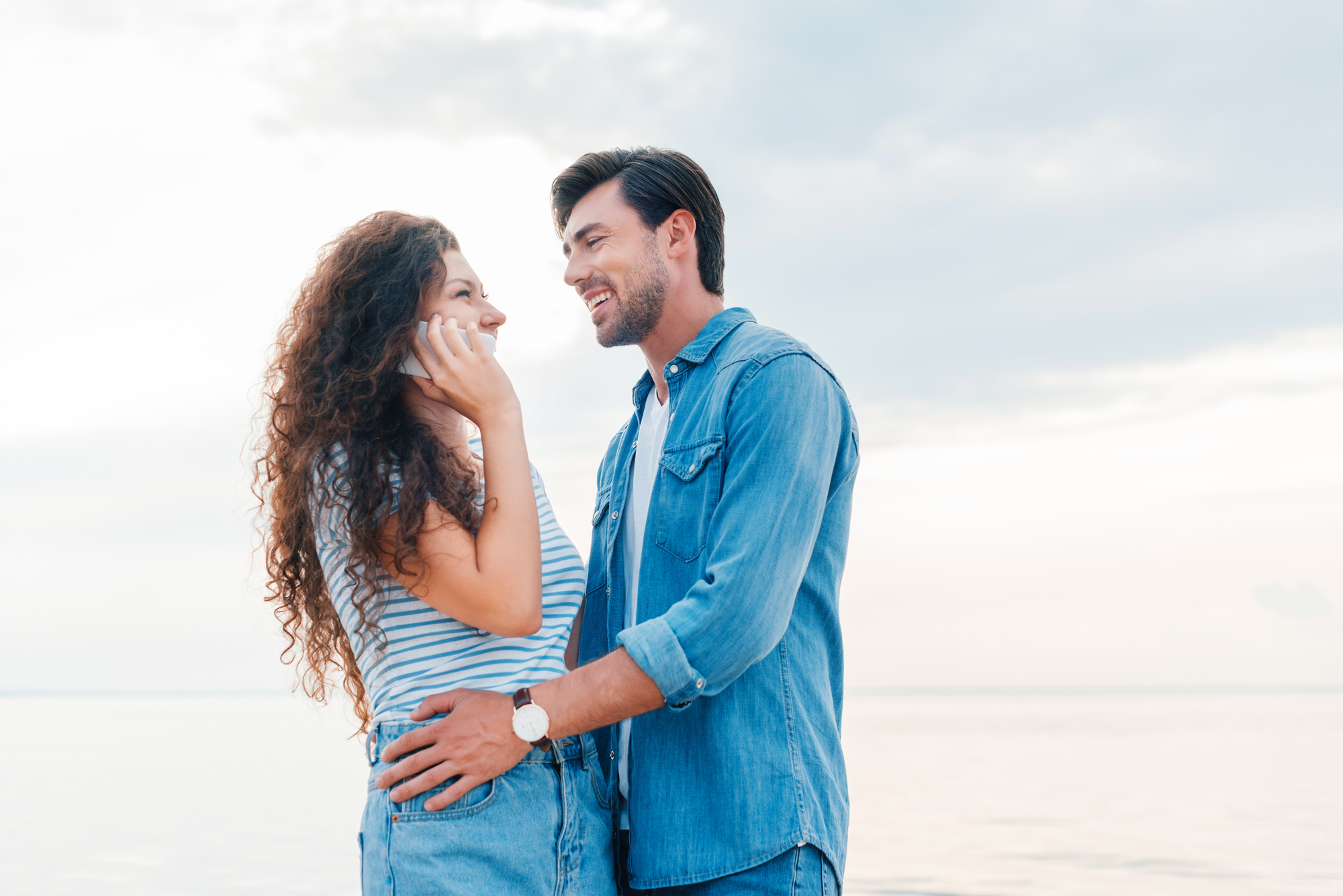 A man and woman stand by a calm body of water under a cloudy sky. The woman has curly hair and is wearing a striped shirt, while the man has short hair and is wearing a denim shirt. They are smiling at each other and appear to be enjoying a conversation.