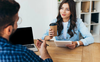 A woman with long brown hair, wearing a denim shirt, is sitting at a wooden table holding a takeaway coffee cup and a tablet, smiling at a man sitting opposite her. The man is in a plaid shirt, holding a pen. A laptop and bookshelf are in the background.