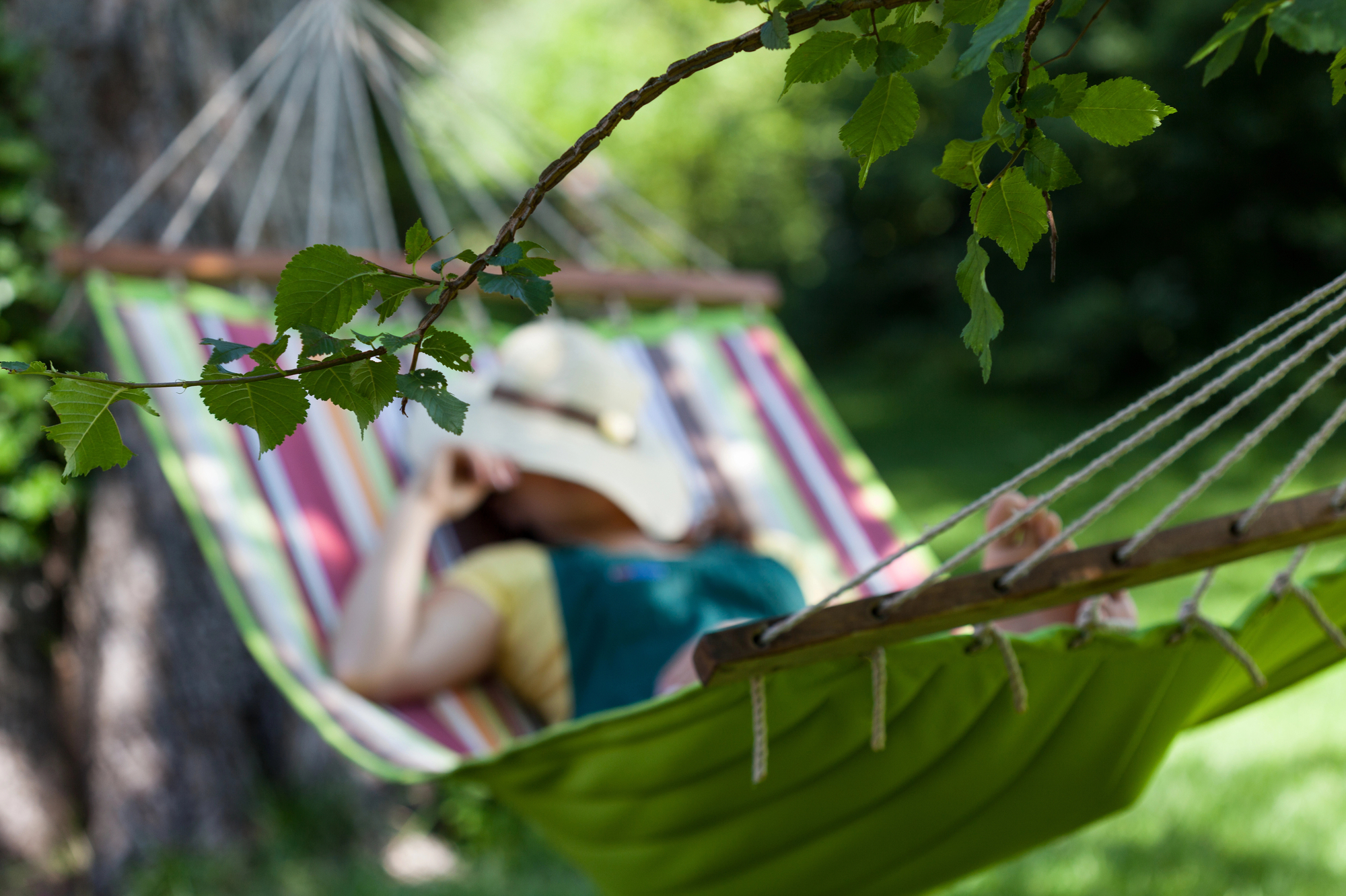 A person wearing a sunhat and casual clothes lounges in a green, striped hammock tied to a tree in a lush garden. They are positioned with their feet up and hands behind their head, creating a relaxed and tranquil atmosphere. Leaves are in focus in the foreground.