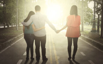 Three people walking down a sunlit road with trees on either side. A man in the middle has his arms around two women, one on his left wearing a teal shirt and the other on his right wearing a pink shirt. They hold hands and face away from the camera.