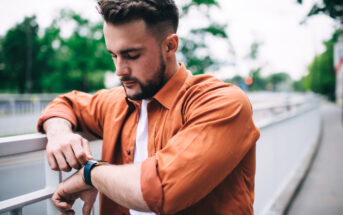 A man with short hair and a beard, dressed in a white t-shirt and an orange button-up shirt, stands outdoors by a railing, looking at his smartwatch. Trees and a pedestrian path are visible in the background.