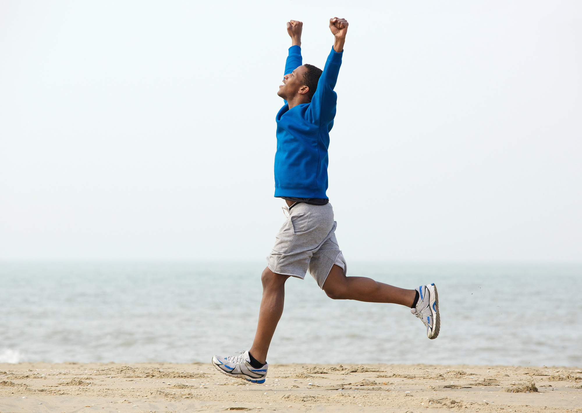 A person wearing a blue sweatshirt, gray shorts, and running shoes is running on a beach with arms raised in the air. The ocean is visible in the background, and the sky is clear. The individual appears joyful and energetic.