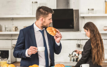 A man in a suit enjoys a croissant and holds a cup of coffee while standing in a modern kitchen. A woman in the background, also dressed in business attire, pours herself a drink from a kettle. Croissants and a plate of fruits are on the counter.