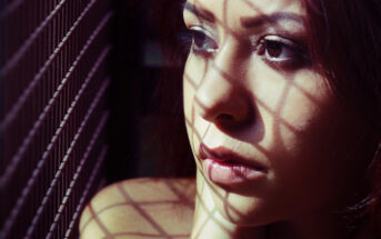 A woman with long hair looks out pensively, with shadows from a grid pattern cast across her face. She appears thoughtful, and the light highlights the intricate shadows, creating a dramatic effect on her expression.