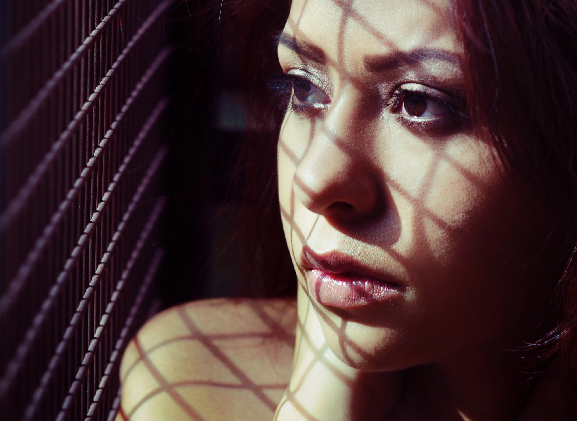 A woman with long hair looks out pensively, with shadows from a grid pattern cast across her face. She appears thoughtful, and the light highlights the intricate shadows, creating a dramatic effect on her expression.