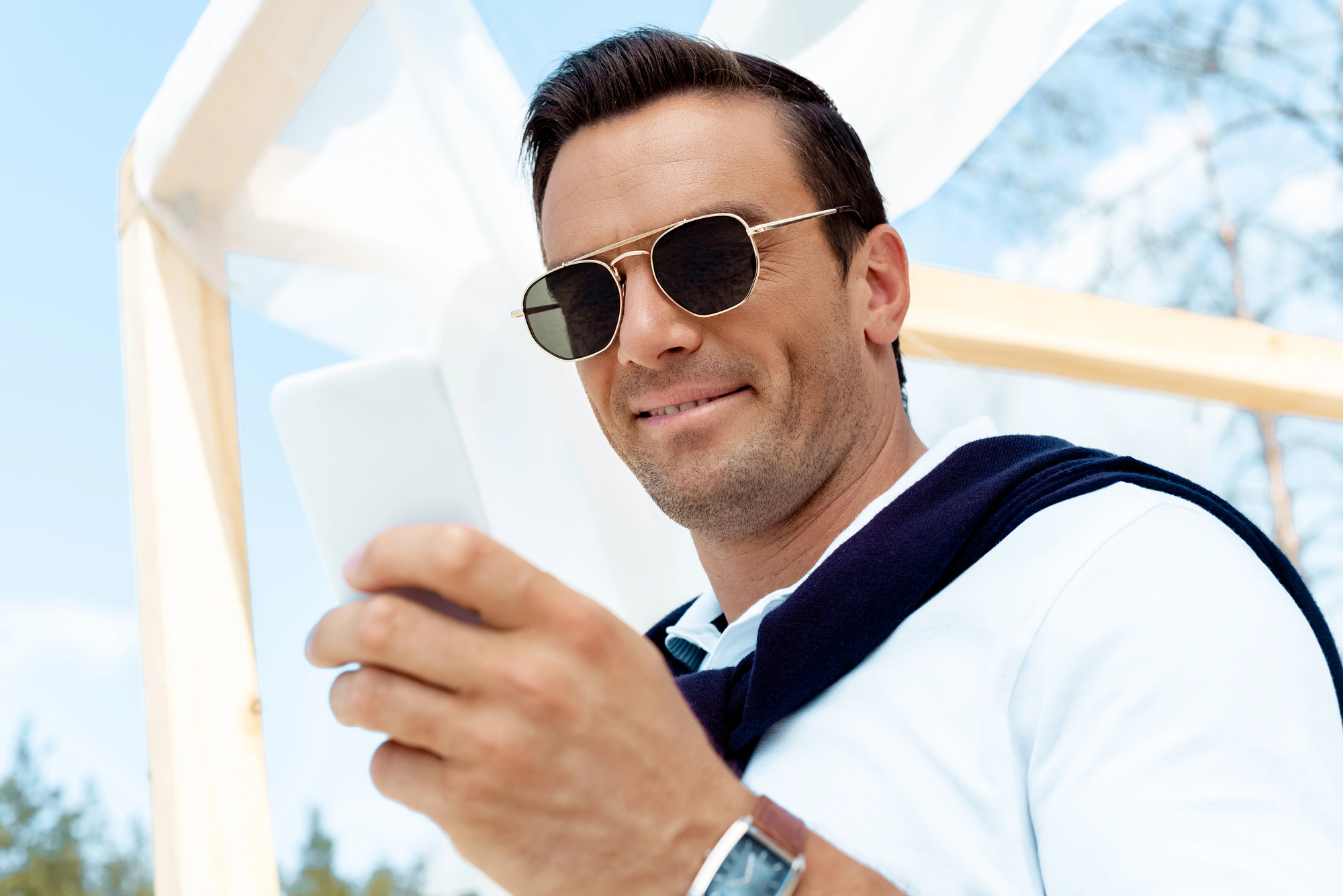 A man wearing sunglasses and a white shirt with a dark sweater draped over his shoulders is smiling while looking at his phone. He has a watch on his wrist and stands outdoors under a light wooden structure with white fabric. Trees and blue sky are in the background.