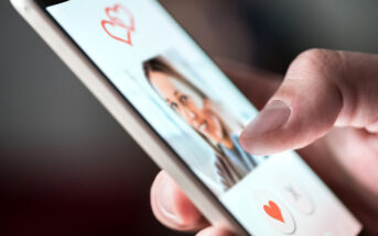 Close-up of a hand holding a smartphone displaying a dating app. A person's profile photo with hearts is visible on the screen, while the thumb is about to swipe right on the heart icon to show interest.