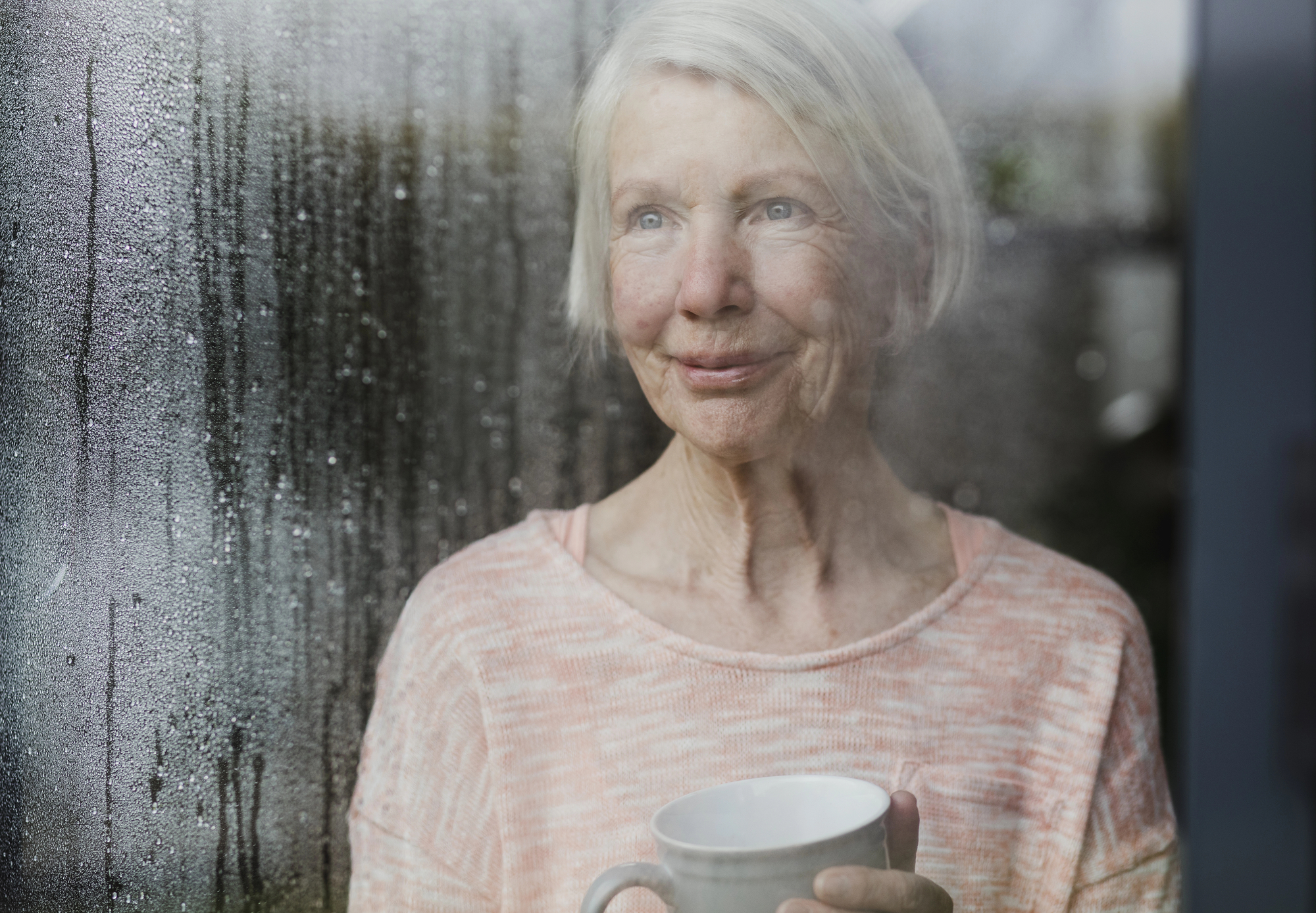 An elderly woman with short gray hair gazes thoughtfully out of a window on a rainy day, holding a white mug. She wears a light pink sweater, and the window is covered with raindrops. The background outside the window is slightly blurred.