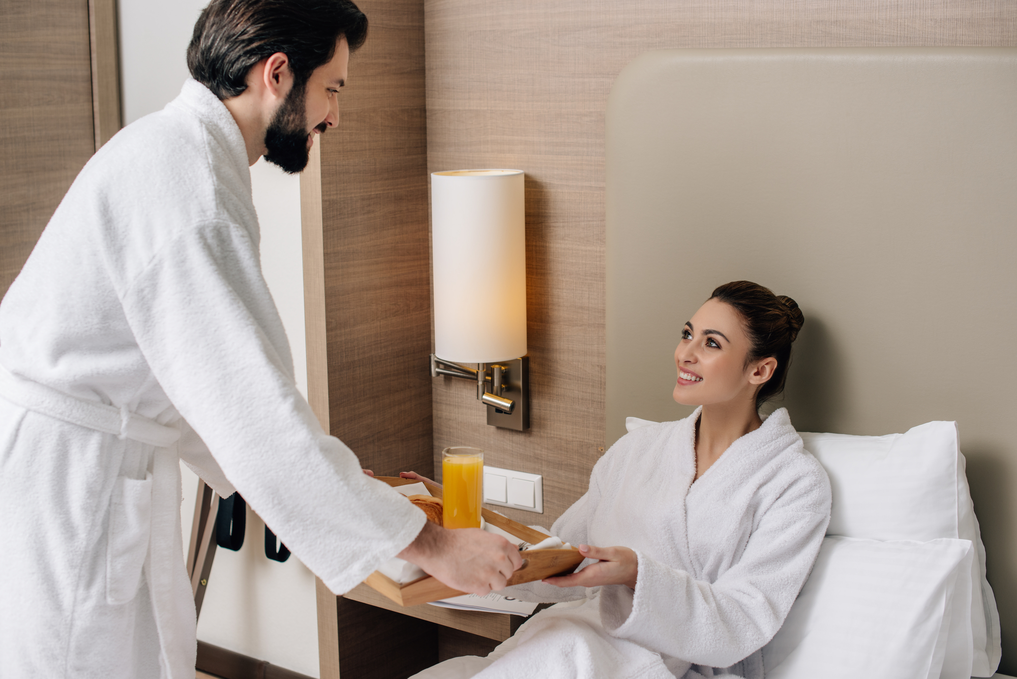 A man in a white bathrobe is serving a woman, also in a white bathrobe, breakfast in bed. The woman is sitting up in bed, smiling, and receiving a tray with food and a glass of orange juice from the man. The setting appears to be a cozy hotel room.