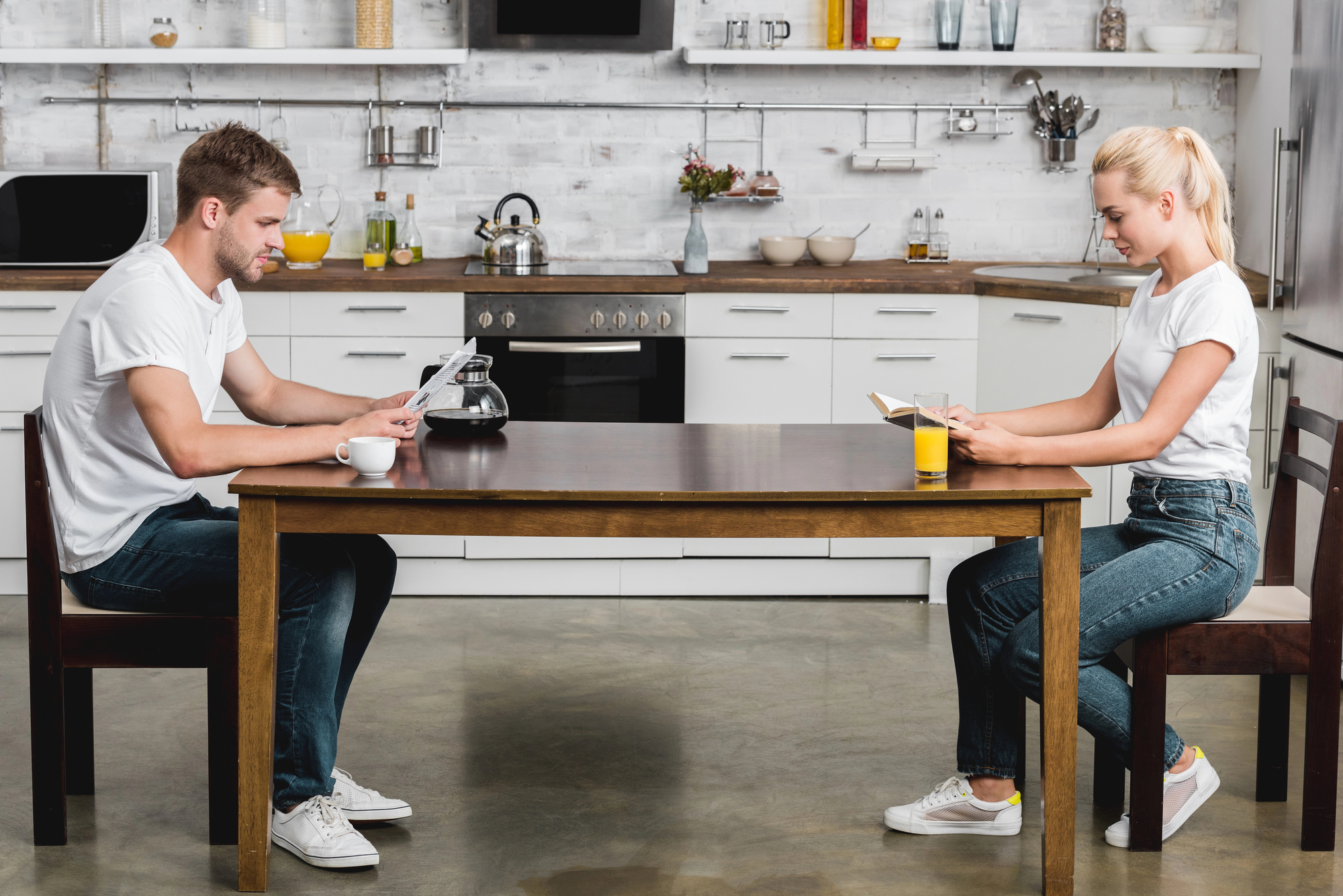 A man and woman sit across from each other at a wooden table in a bright kitchen, both focused on their respective tablets. They are dressed casually in white t-shirts and jeans. The kitchen features white cabinets, a microwave, a stovetop, and various kitchen items.