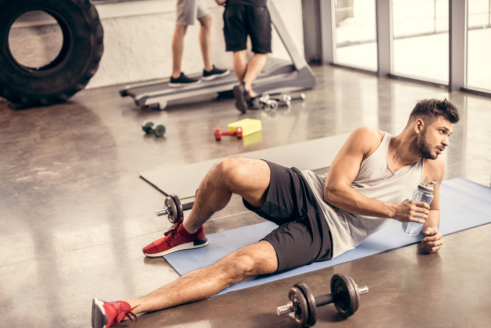 A man in athletic wear relaxes on a gym mat while holding a water bottle. Dumbbells are scattered nearby on the floor. In the background, two other individuals are working out on a treadmill near large windows. A large tire is also visible against the wall.