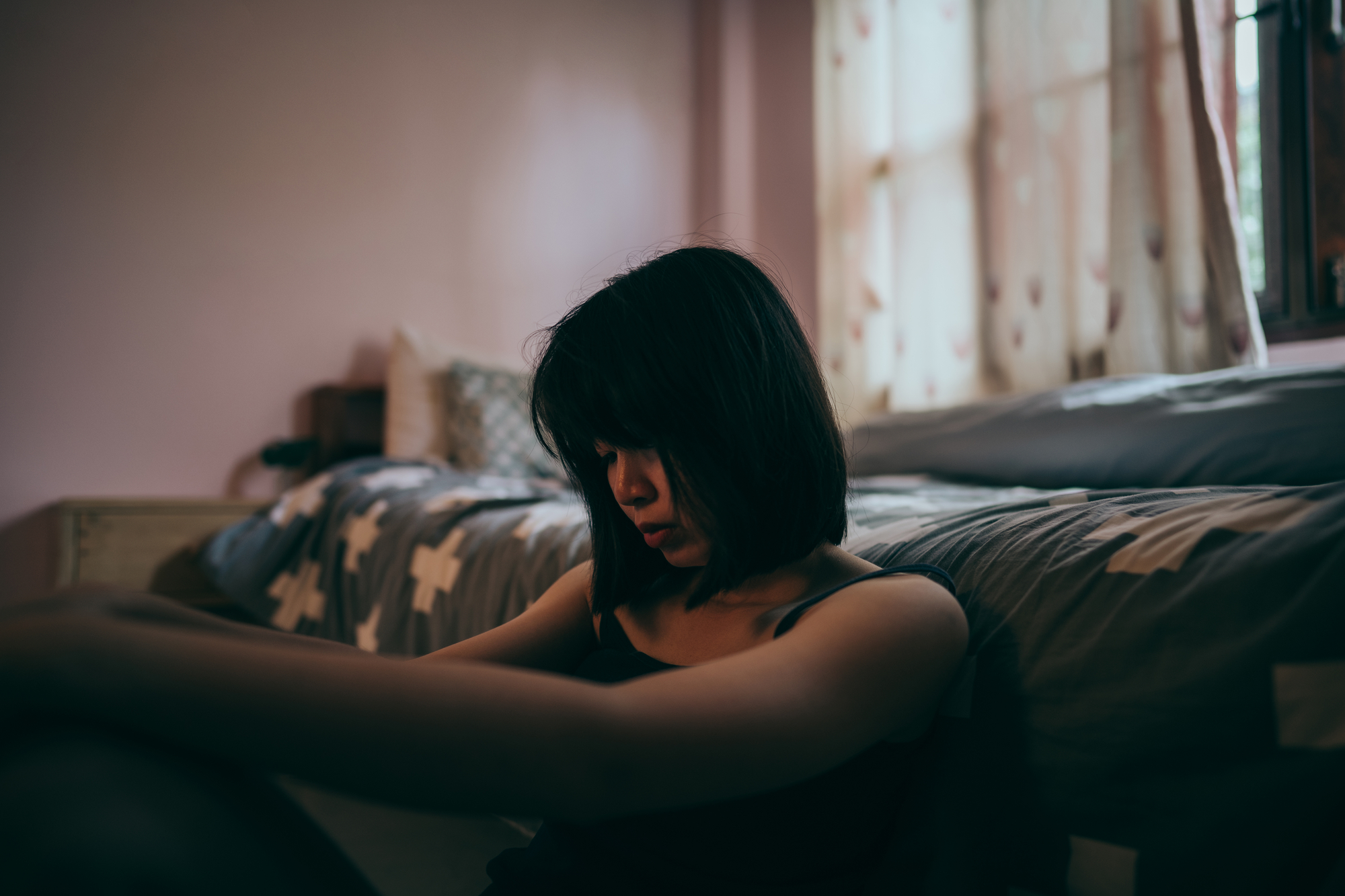 A young woman with dark hair sits on the floor against a bed, looking down with a contemplative or somber expression in a dimly lit bedroom with light coming through the window. The room has soft tones, with curtains and bedspreads in muted colors.