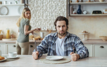 A man in a plaid shirt sits at a dining table with an empty plate, holding a fork and knife, looking frustrated. In the background, a woman in a green shirt prepares food at the kitchen counter and looks back towards the man. The kitchen is modern and well-lit.