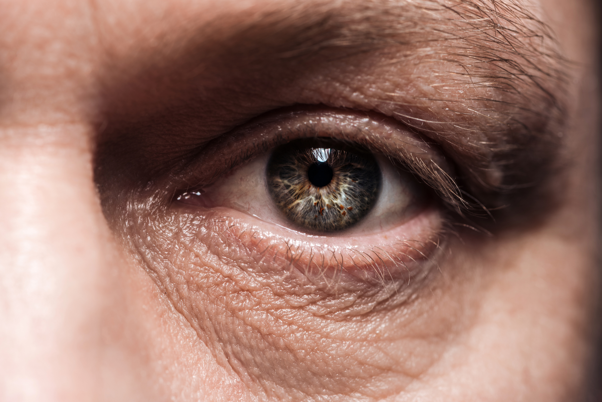 Close-up image of a person's eye. The eye is hazel with intricate detail and reflections visible on the iris. The surrounding skin shows natural texture with fine lines and wrinkles, indicating an expressive face. The eyelid and eyebrow are partially visible.