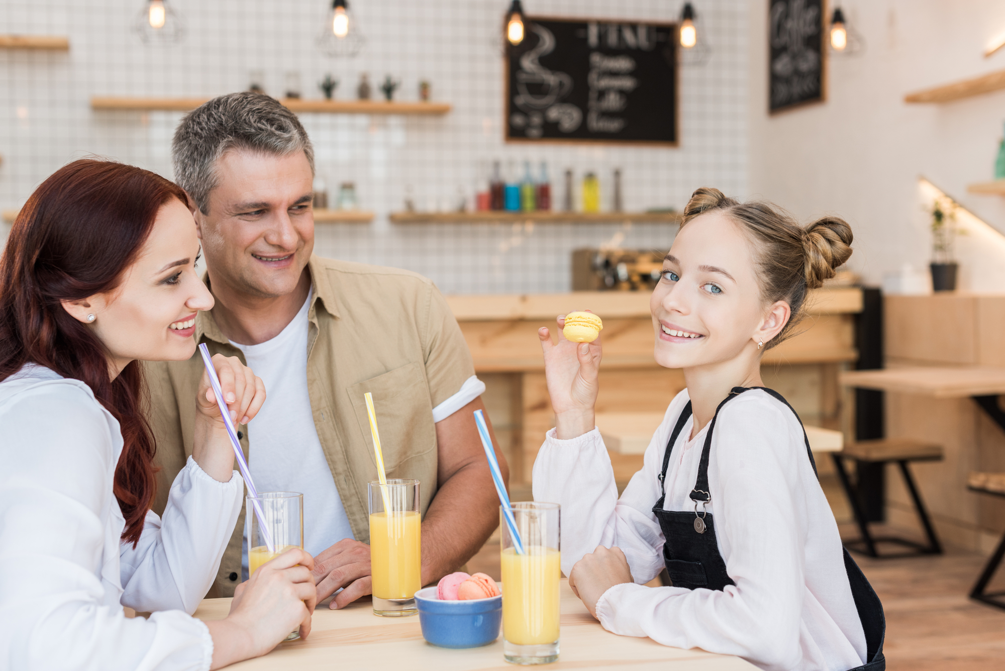 A family of three, comprising a woman, a man, and a young girl with buns in her hair, sit at a café table. They smile and enjoy glasses of orange juice with straws. The girl holds a yellow macaron, and a bowl of macarons sits on the table. The background is a trendy café.