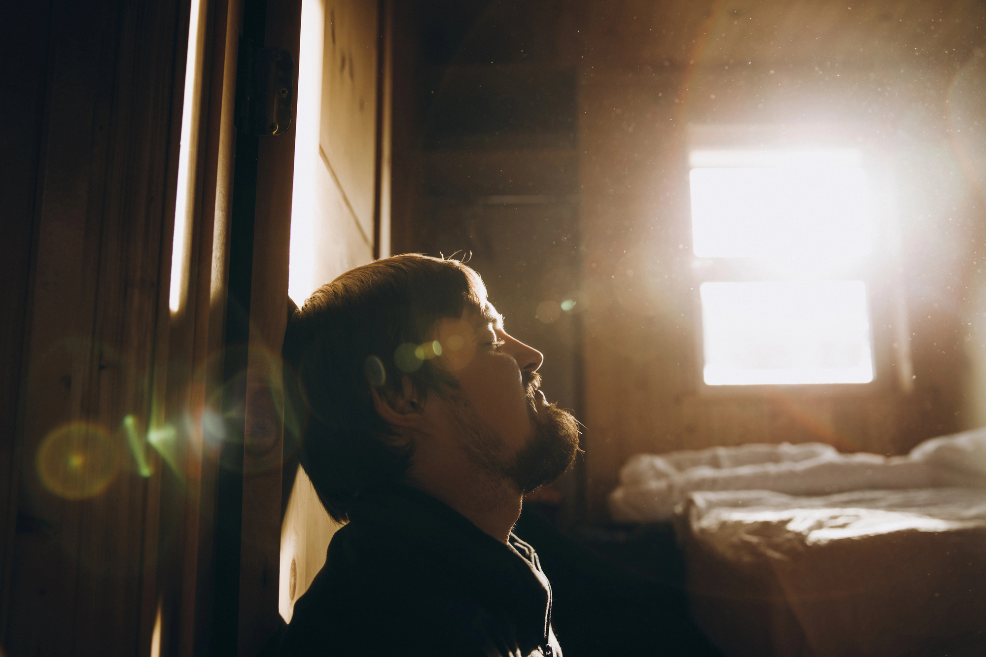 A person with a beard leans against a door, bathed in warm sunlight streaming through a window in a cozy, rustic room. Dust particles are visible in the golden light, and an unmade bed is in the background, creating a serene and contemplative atmosphere.