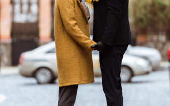 Two people standing face-to-face outdoors, holding hands. One wears a mustard-yellow coat, while the other wears a dark coat and black gloves. Cars are blurred in the background on the cobblestone street.