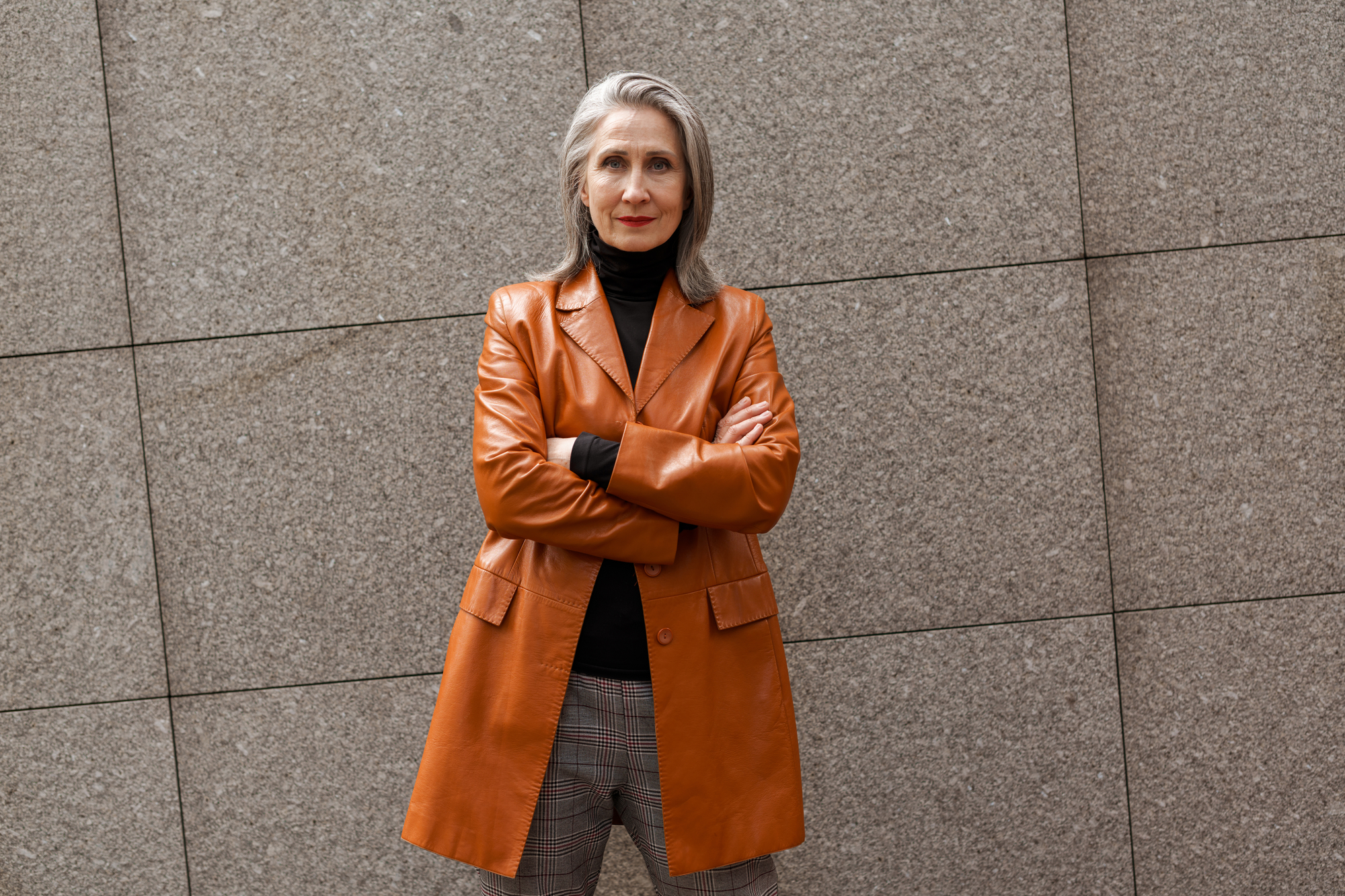 A confident woman with long gray hair stands against a textured stone wall. She is wearing a brown leather jacket over a black turtleneck and plaid pants, with her arms crossed and a serious expression on her face.