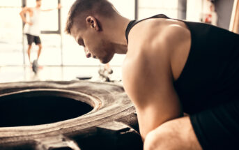 A person wearing a black tank top is focused on lifting a large tire in a gym. In the background, another person can be seen standing with one arm resting on a window frame, watching. The gym has bright natural light coming through the windows.