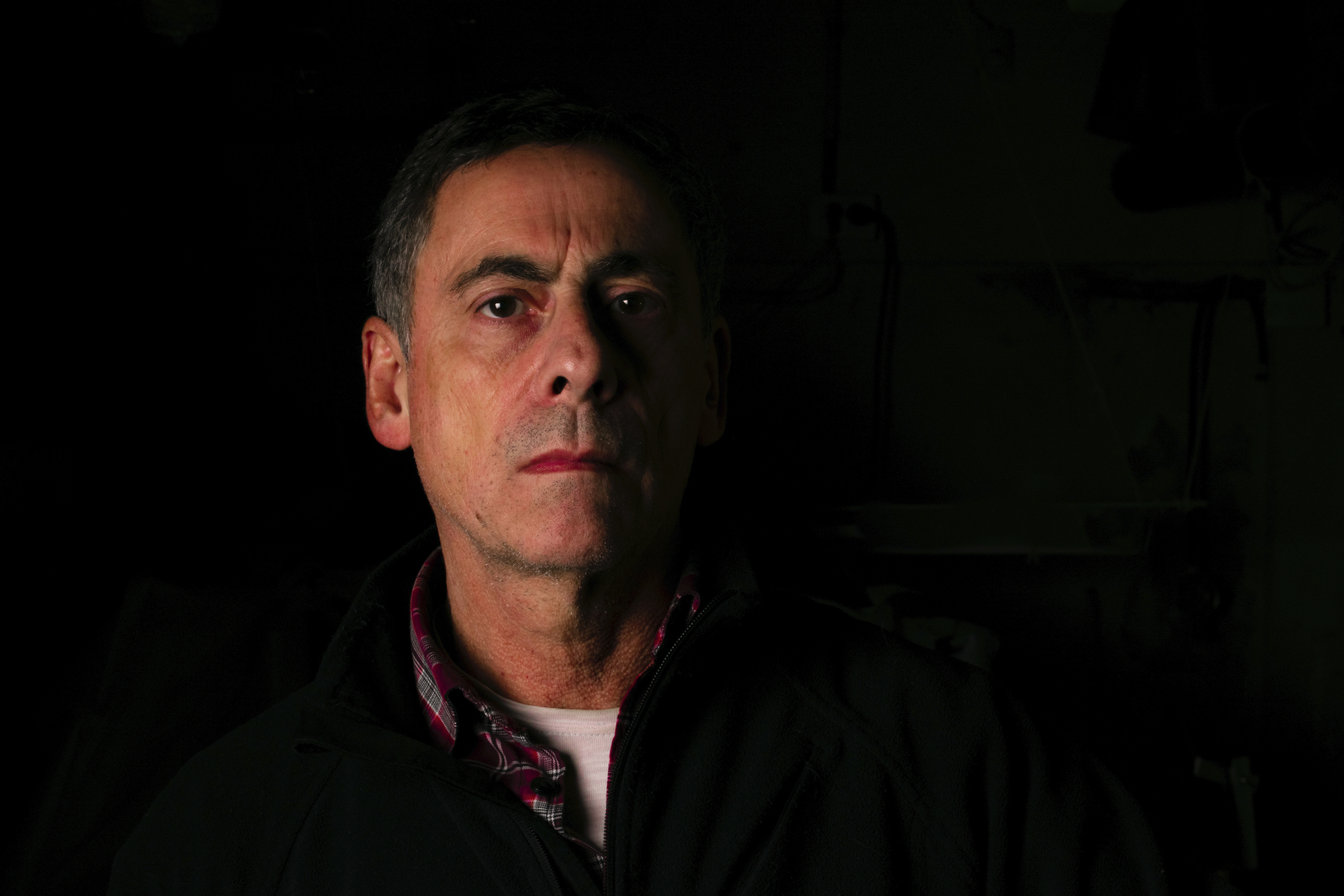 A middle-aged man with graying hair and a serious expression on his face stands in a dimly lit environment. He is wearing a dark jacket over a checkered shirt, with shadowed surroundings that make his face the primary focus.