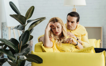 A woman, looking distressed with her hand on her forehead, sits on a yellow couch. A man beside her rests his arm on her shoulders, providing comfort. Both are wearing yellow tops. A large leafy plant is in the foreground, and a lamp is in the background.