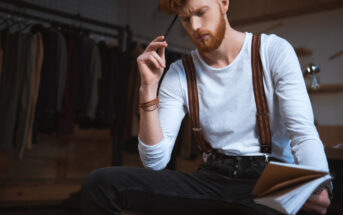 A bearded man with red hair, wearing a white shirt and suspenders, sits pensively holding a pen to his mouth. He is looking at a notebook in his hand. In the background, there are clothes hanging on a rack, suggesting a fashion or creative workspace.