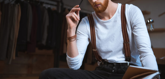 A bearded man with red hair, wearing a white shirt and suspenders, sits pensively holding a pen to his mouth. He is looking at a notebook in his hand. In the background, there are clothes hanging on a rack, suggesting a fashion or creative workspace.