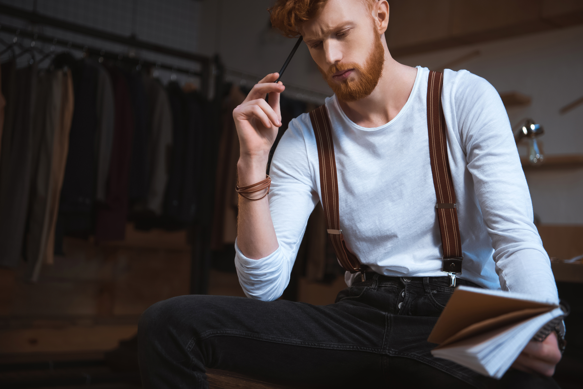 A bearded man with red hair, wearing a white shirt and suspenders, sits pensively holding a pen to his mouth. He is looking at a notebook in his hand. In the background, there are clothes hanging on a rack, suggesting a fashion or creative workspace.