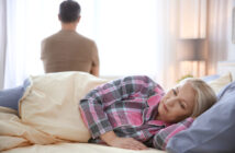 A woman lying in bed, wearing plaid pajamas, looking pensive. In the background, a man in a brown shirt sits at the edge of the bed, facing away and looking toward a bright window. The room is softly lit with a table lamp on a nightstand.