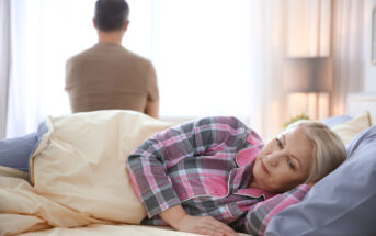 A woman lying in bed, wearing plaid pajamas, looking pensive. In the background, a man in a brown shirt sits at the edge of the bed, facing away and looking toward a bright window. The room is softly lit with a table lamp on a nightstand.