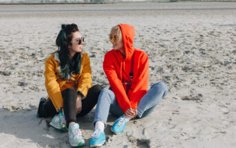 Two people sit on a sandy beach, facing each other and smiling. One wears a yellow jacket, black pants, and has dark hair, while the other wears a bright red hoodie, blue jeans, and has blond hair. The background features a wide expanse of sand and a distant horizon.