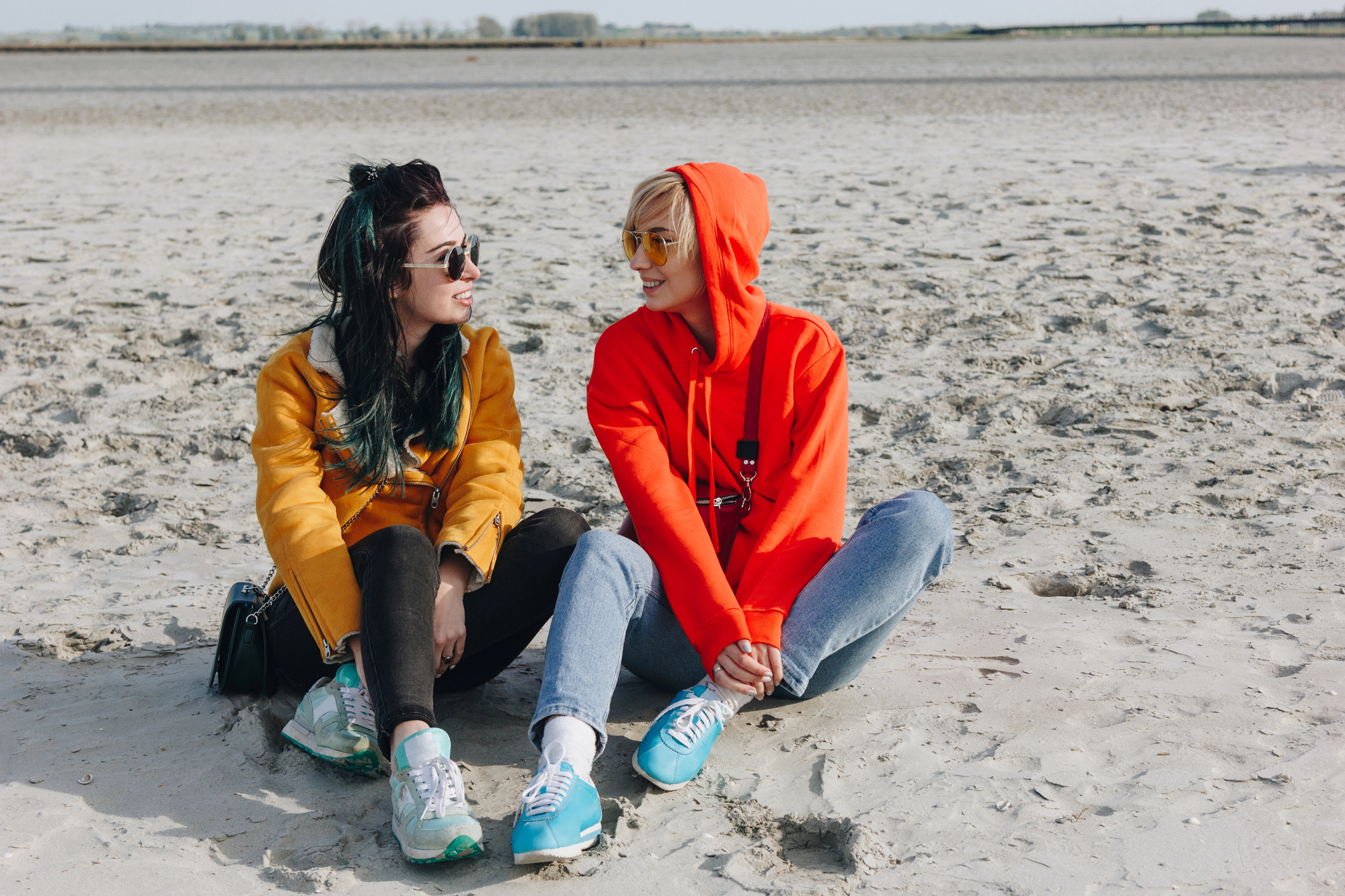 Two people sit on a sandy beach, facing each other and smiling. One wears a yellow jacket, black pants, and has dark hair, while the other wears a bright red hoodie, blue jeans, and has blond hair. The background features a wide expanse of sand and a distant horizon.