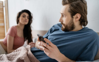 A man with a beard is lying in bed, holding a smartphone and looking away with a concerned expression. A woman with long dark hair and wearing a pink top sits beside him, looking at him with a serious expression. They're covered with a light pink blanket.