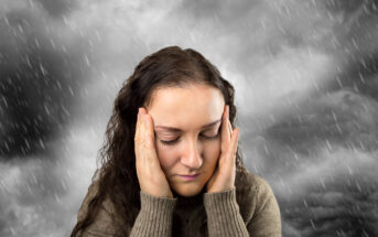 A woman with closed eyes and a distressed expression holds her hands to her head. She is wearing a brown sweater. Behind her, a dramatic black-and-white stormy sky with heavy rain emphasizes the intensity of her emotion.