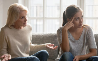 A senior woman and a younger woman sit on a couch in front of a window. The older woman appears to be explaining something with an expressive gesture while the younger woman, sitting with her hand on her forehead, looks away with a frustrated expression.