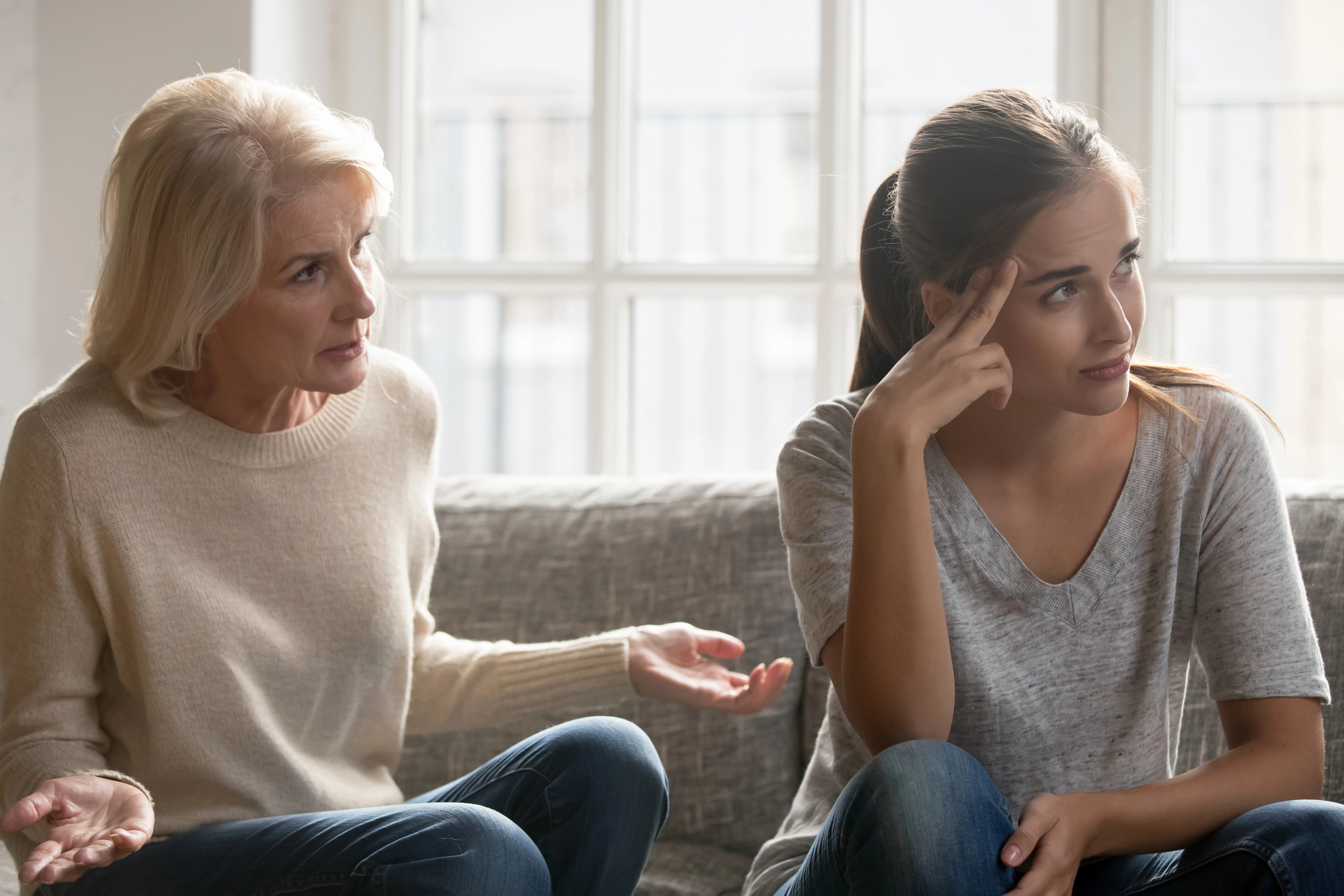 A senior woman and a younger woman sit on a couch in front of a window. The older woman appears to be explaining something with an expressive gesture while the younger woman, sitting with her hand on her forehead, looks away with a frustrated expression.