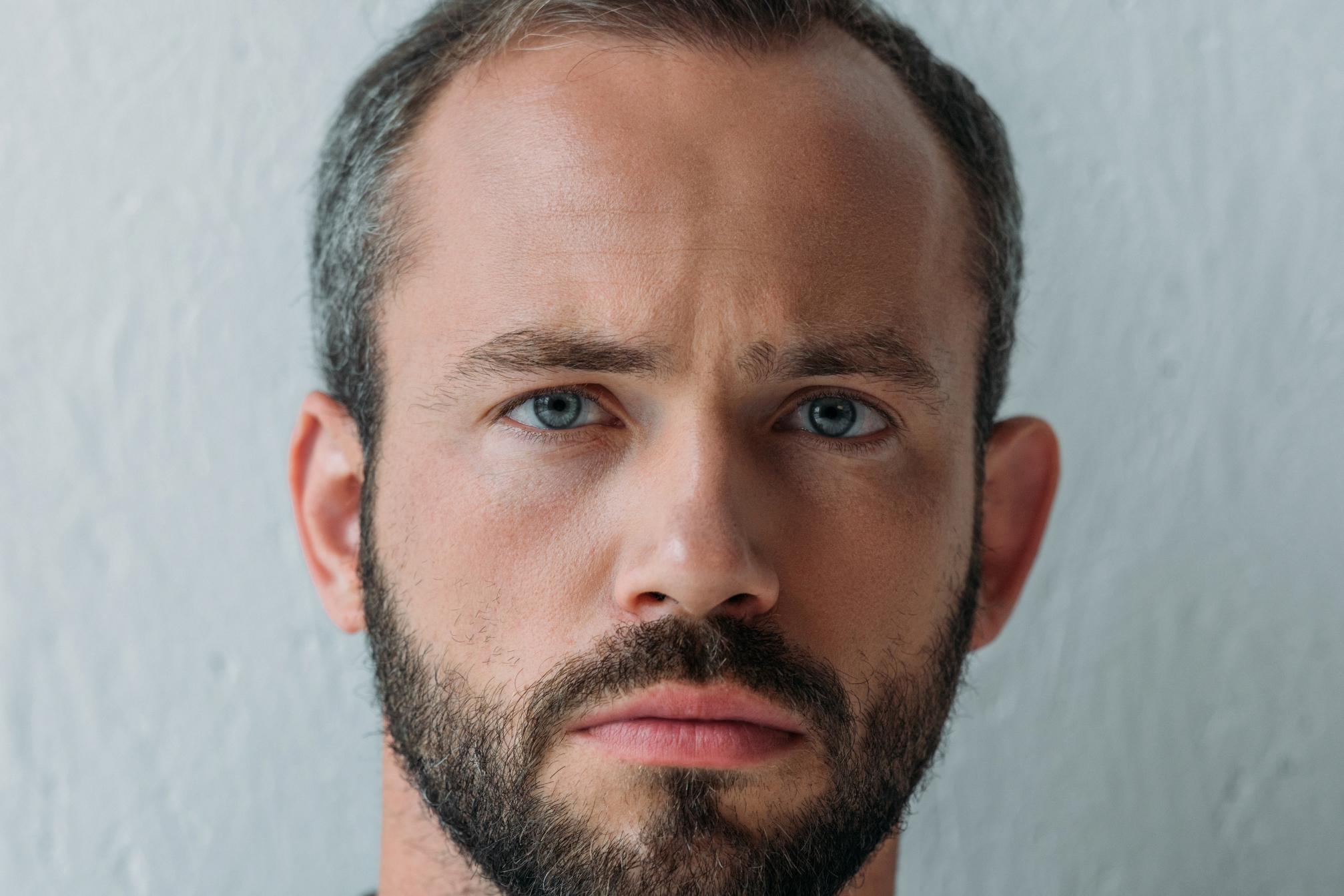 A man with short brown hair and a full beard stands against a light-colored, textured wall. He has blue eyes and is looking straight at the camera with a neutral expression.