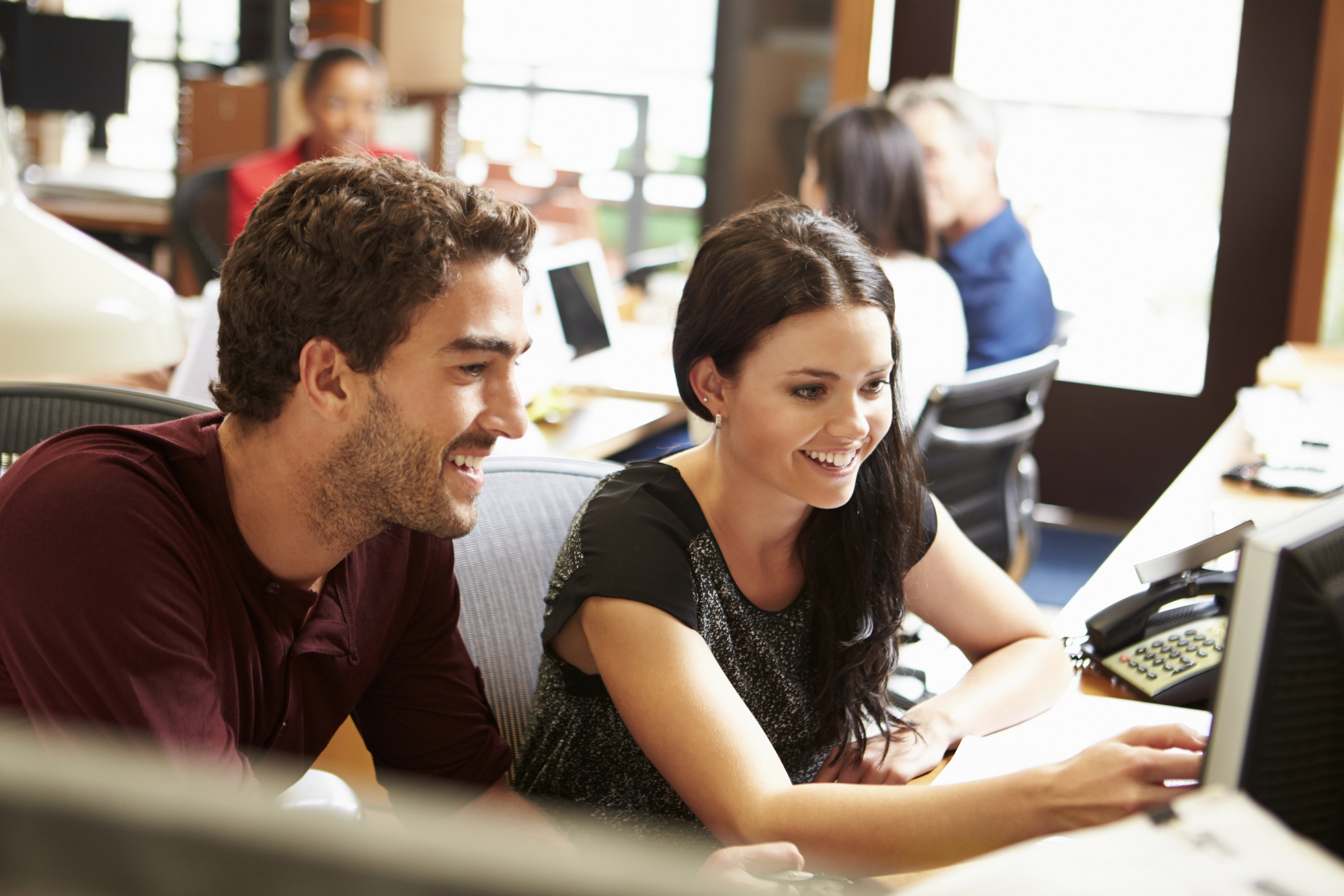 A man and a woman are seated at a desk in an office, smiling as they look at a computer screen. The office is bright with natural light, and other colleagues are visible working in the background. Both appear engaged and collaborative in their work.