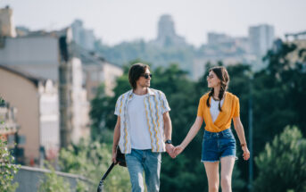 A man and a woman are walking hand in hand outdoors. The man, holding a guitar, is wearing sunglasses, a white shirt with a yellow pattern, and blue jeans. The woman, also in sunglasses, is dressed in a yellow top and denim shorts. The background shows city buildings and greenery.
