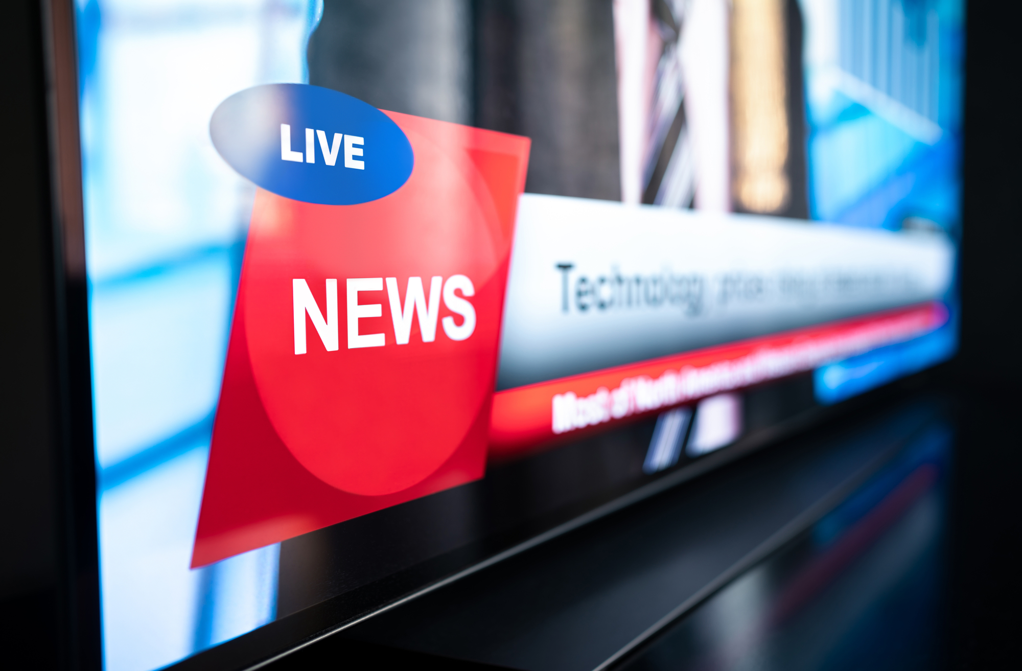 A close-up image of a television screen displaying a live news broadcast. The screen shows the word "NEWS" in large white letters on a red background, with the word "LIVE" in smaller blue letters above it. Text on the screen is partially blurred.