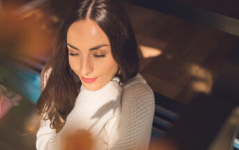 A woman with long brown hair is sitting and facing slightly downward with her eyes closed. She is wearing a white sweater, and sunlight creates soft shadows around her. The setting appears cozy and warm, with blurred elements in the foreground.