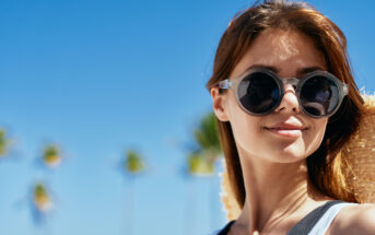 A young woman with long brown hair wearing round sunglasses and a light-colored top smiles under a clear blue sky. Palm trees are blurred in the background, creating a sunny, tropical atmosphere.