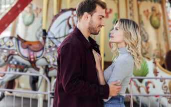 A couple stands facing each other in front of a colorful carousel, with the woman resting a hand on the man's chest. The man wears a burgundy jacket, and the woman wears a gray long-sleeve top and blue jeans. Both are gazing into each other's eyes.