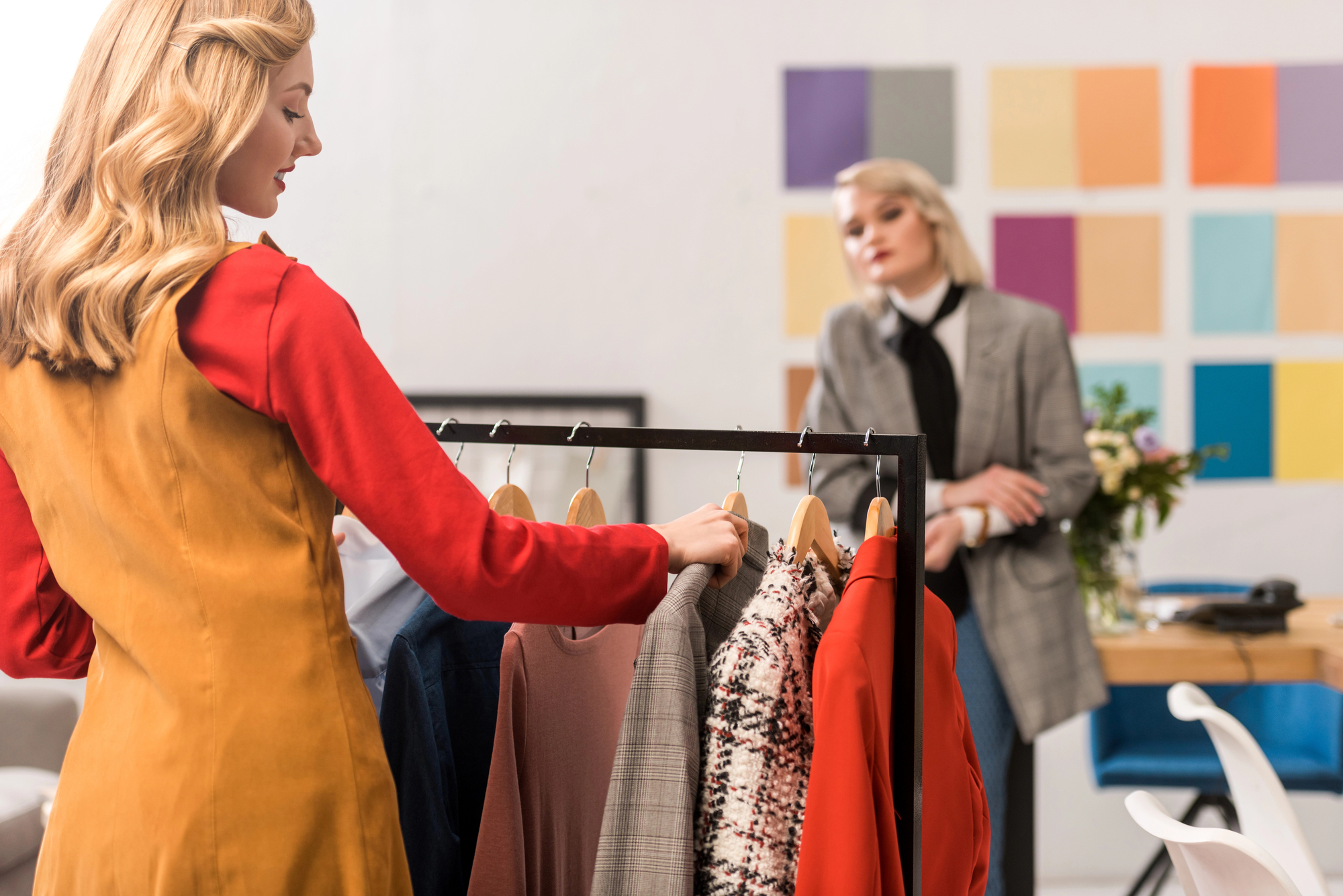 Two women in a room with a colorful wall grid: one woman with blonde hair examines clothes on a hanging rack, while the other, also blonde, sits at a desk with flowers and a laptop. The clothing includes various colors and patterns.